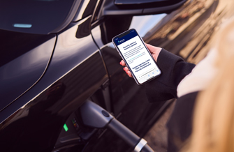 Person holding a cell phone open to the electrify america site next to a vehicle charging.